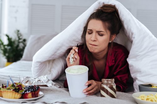 woman lying in bed with covers over her head eating ice cream and sweets after breakup