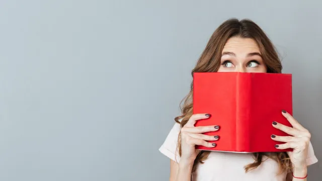 Portrait of a pretty young girl hiding behind an open book and looking away isolated over gray wall background