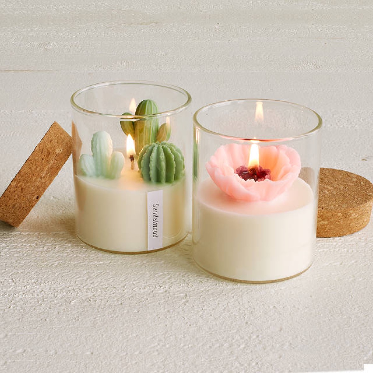 two white candles in glass jars, one with fake cacti and one with a pink flower