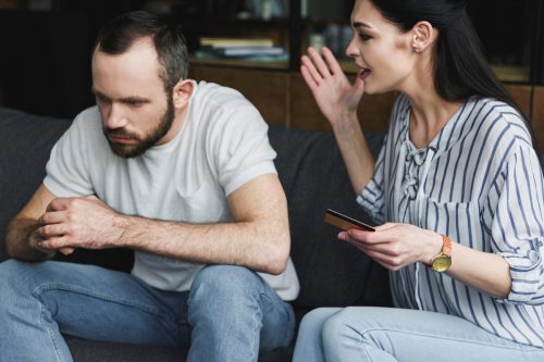 depressed man sitting on couch and looking away while his wife shouts at him holding credit card
