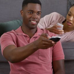 Black man watching TV while woman sits on the couch looking bored