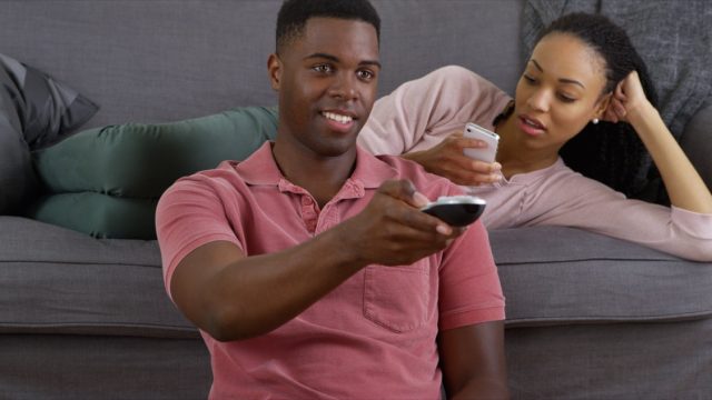Black man watching TV while woman sits on the couch looking bored