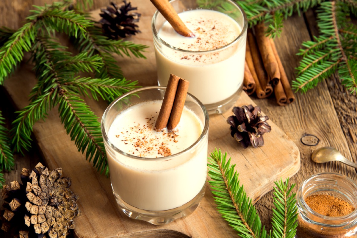 two glasses of egg nog and pine branches on wooden cutting board with pine cones
