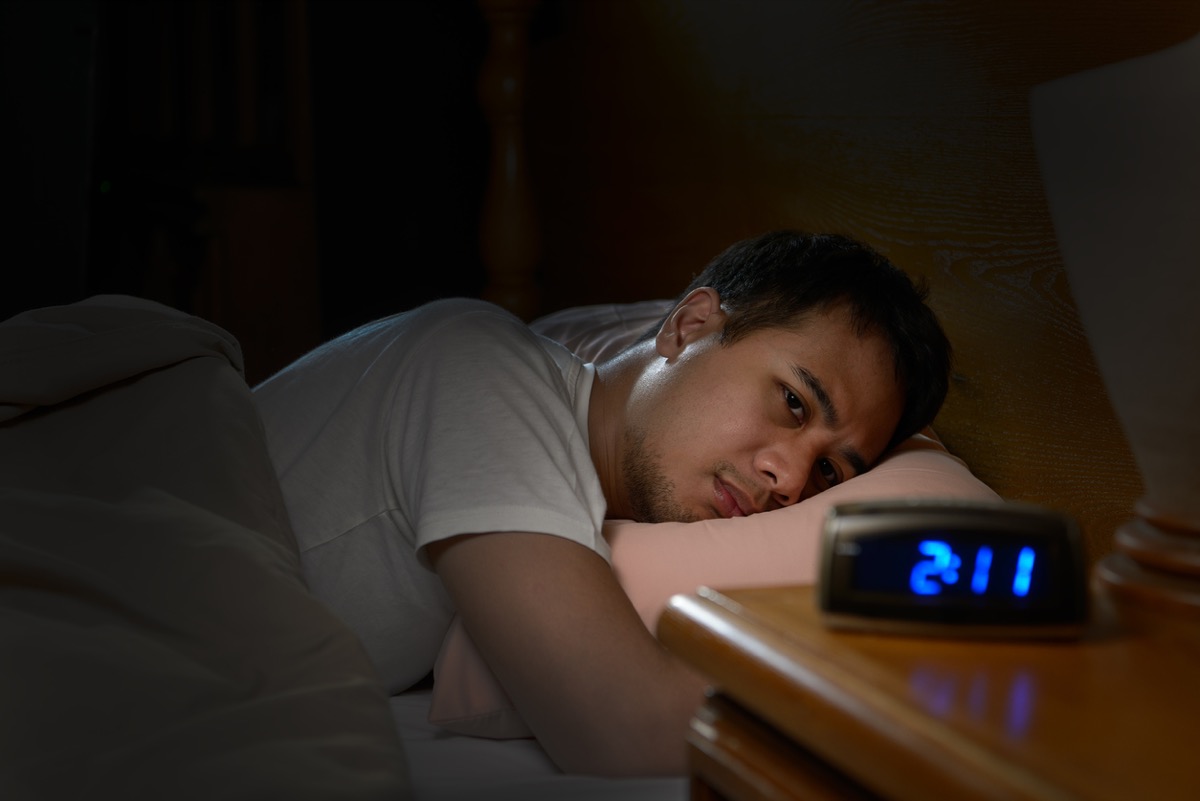 Man suffering from insomnia lying in bed, can't sleep at 2 am, according to clock on nightstand