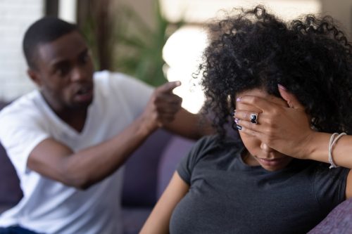 Tired frustrated black woman ignoring angry husband who is pointing his finger at her while she covers her face on the couch