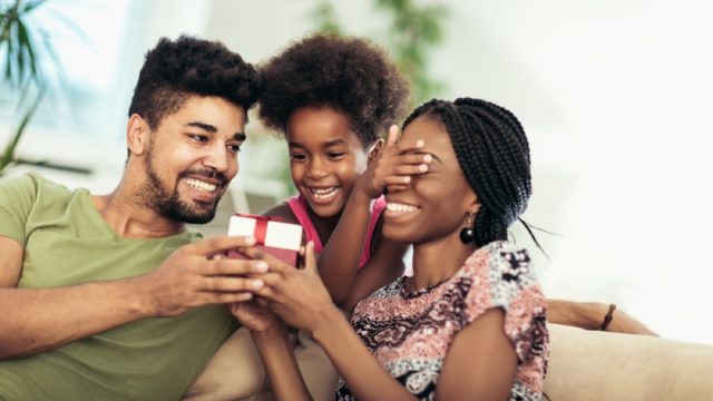 black mother, father, and daughter opening present on couch