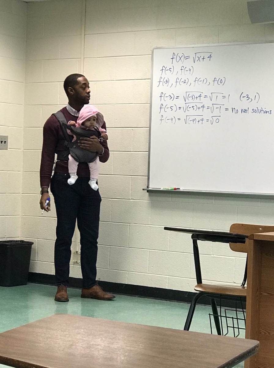 black professor standing in front of white board in classroom with baby in carrier on his chest