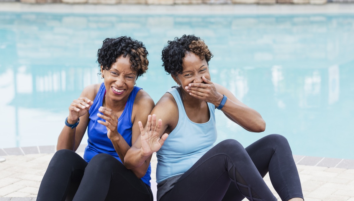 Two mature African-American women in their 40s, identical twins, sitting on a pool deck, laughing uncontrollably, looking at the camera. One of them is holding one hand up in a stop gesture and is covering her mouth with the other as she laughs.