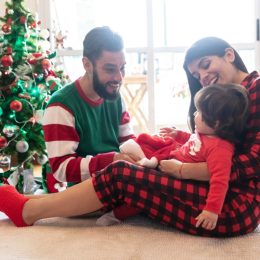 hispanic mother, father, and daughter wearing christmas pajamas in front of christmas tree