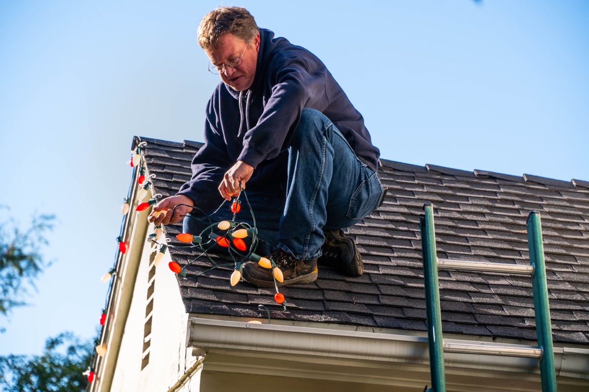 Middle aged man on the rooftop of a house installing a string o Christmas lights. His ladder is nearby.