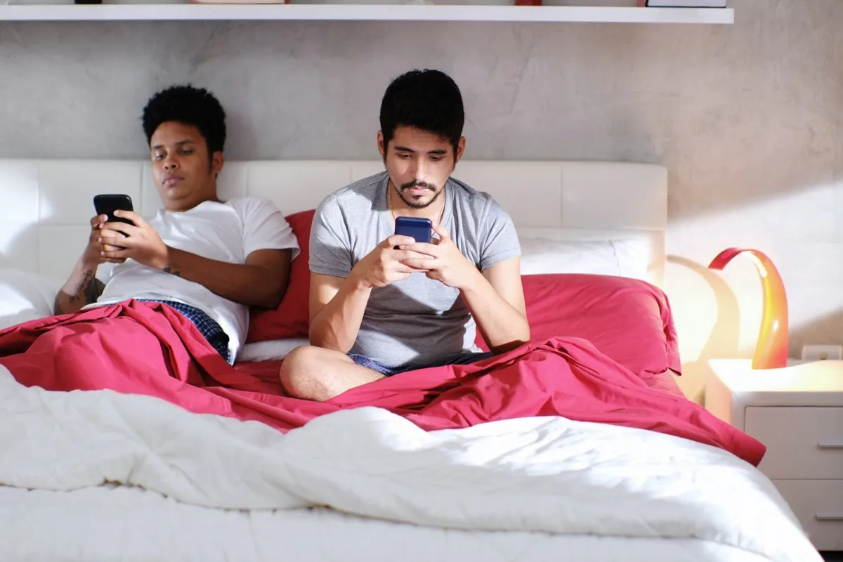 young black man and young asian man in bed together but looking at their phones
