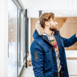 young white man in a winter coat adjusting the thermostat in his home