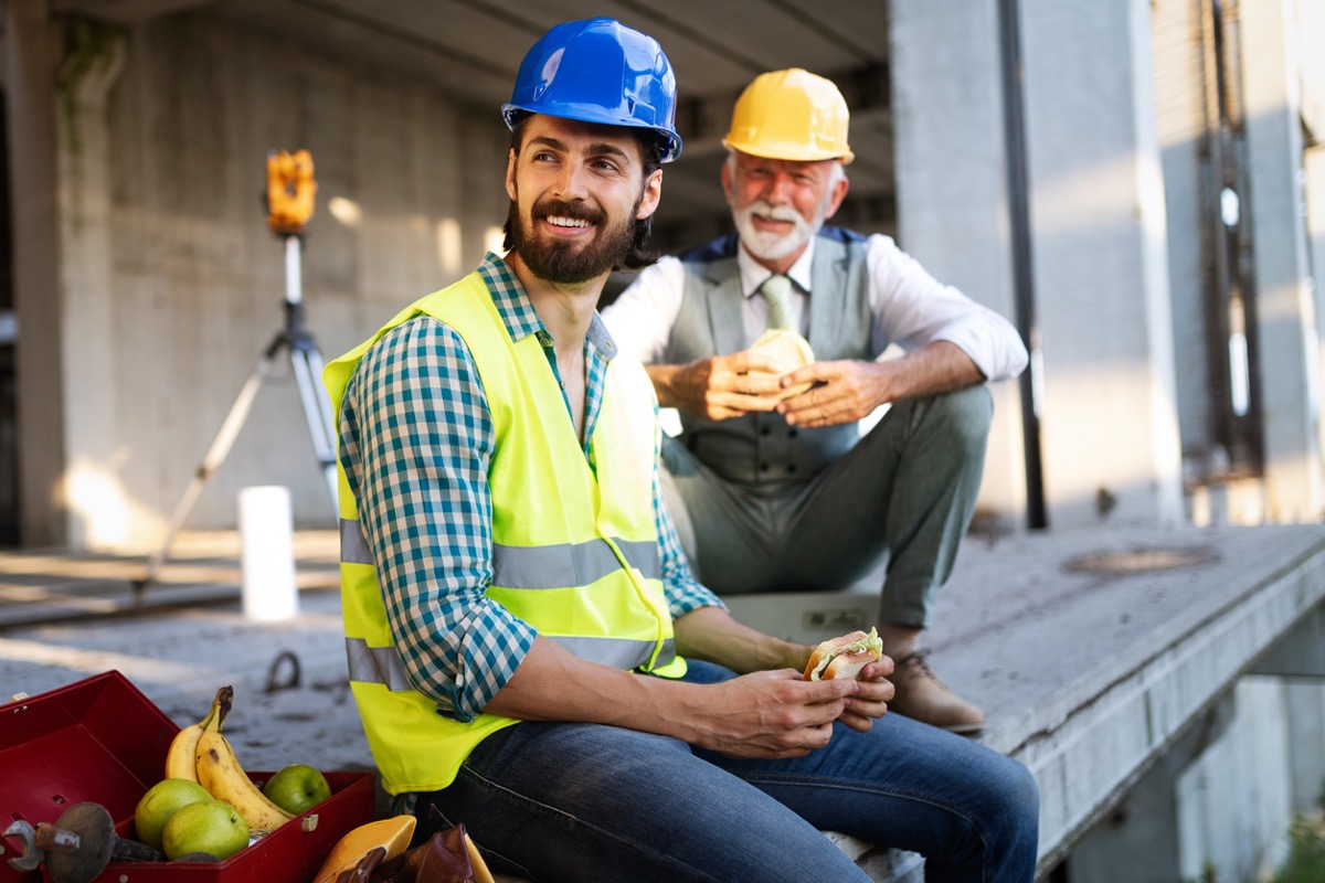 younger and older construction worker eating sandwiches and smiling