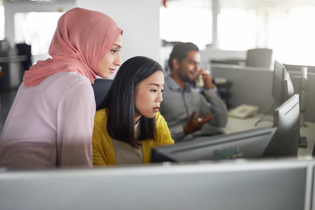 asian woman and muslim woman looking at computer together in an office