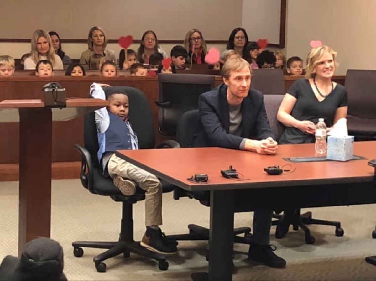 young boy sitting at table in adoption court with his parents and classmates behind him