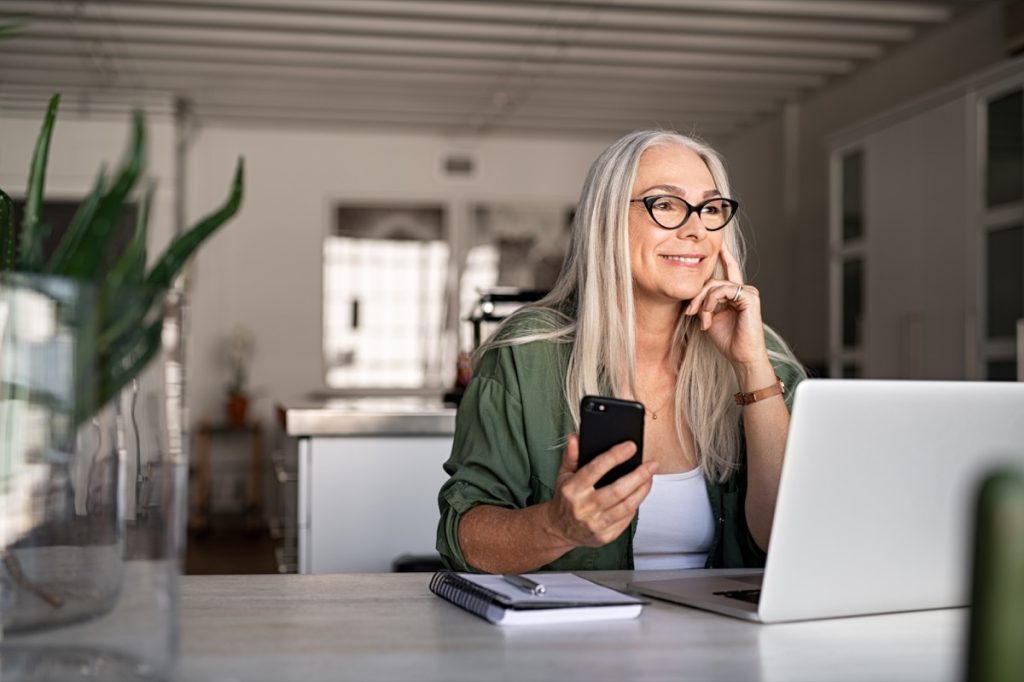 Happy senior woman holding smartphone and laptop daydreaming while looking away. Successful stylish old woman working at home while thinking about a good future. Cheerful fashionable lady entrepreneur wearing cool eyeglasses.