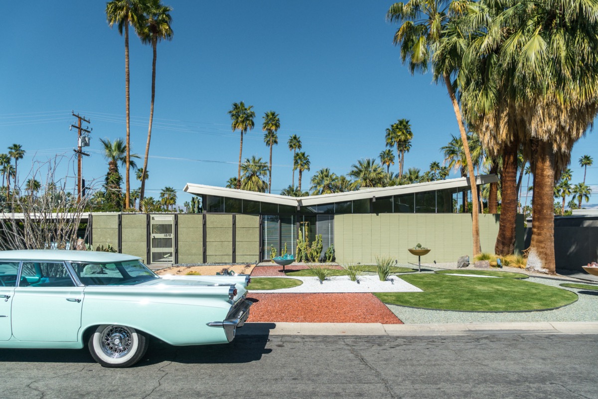 O mașină albastră din anii 1950, parcată în fața unei case moderne de la mijlocul secolului, cu palmieri