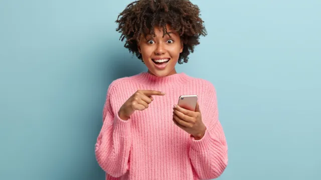 Surprised positive dark skinned lady points at smart phone device, suggests to have conversation in group chat, glad to recieve many messages of congratulation on mailbox, has overjoyed look