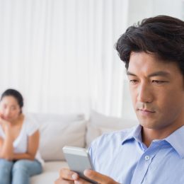 asian man on phone while upset wife looks on from bed