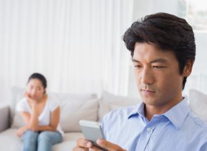 asian man on phone while upset wife looks on from bed