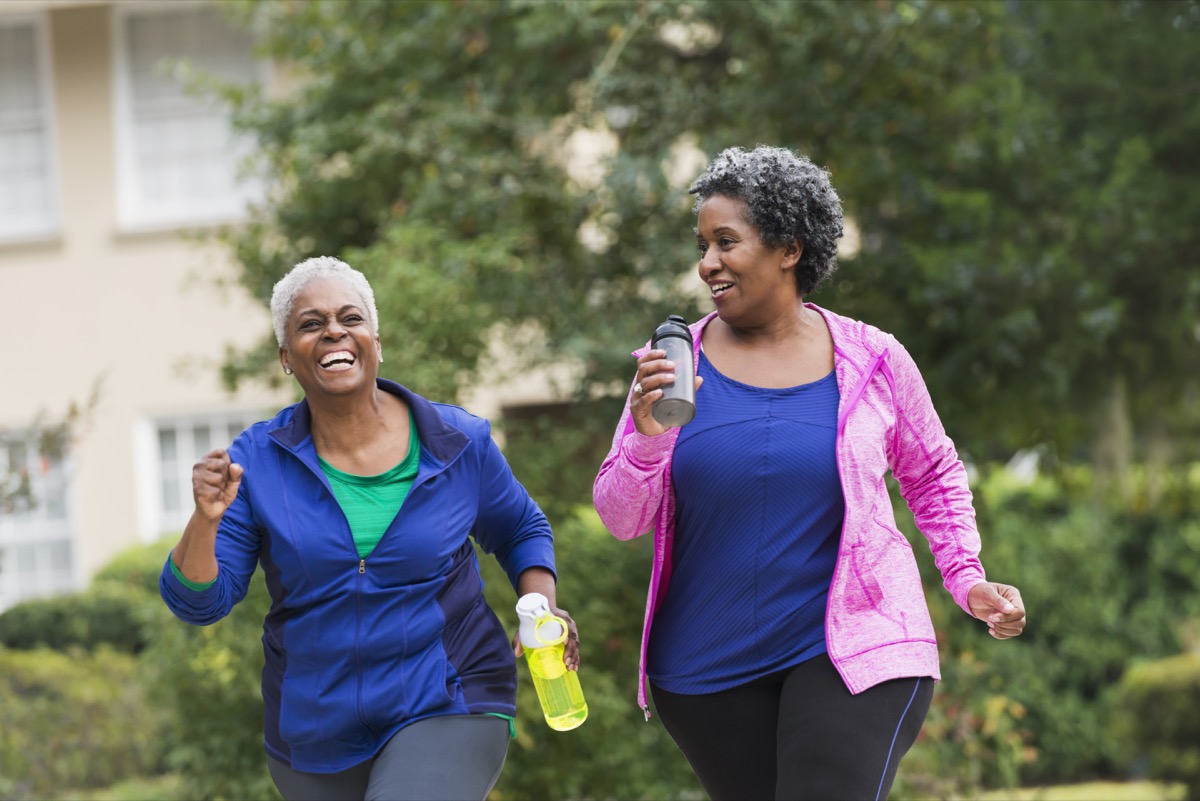 Two senior African American women getting in shape together. They are jogging or power walking on a sidewalk in a residential neighborhood, talking and laughing.