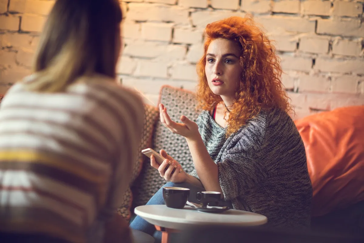 young red-haired woman sitting in a cafe with friend and talking about something.