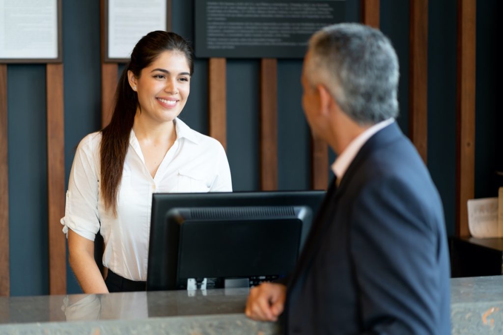  hotel receptionist standing behind counter talking to unrecognizable male guest smiling