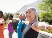 Portrait of happy senior woman practicing yoga outdoor with fitness class. Beautiful mature woman stretching her arms and looking at camera outdoor. Portrait of smiling serene lady with outstretched arms at park. (Portrait of happy senior woman pract