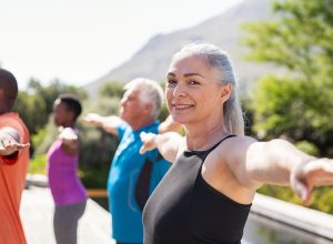 Portrait of happy senior woman practicing yoga outdoor with fitness class. Beautiful mature woman stretching her arms and looking at camera outdoor. Portrait of smiling serene lady with outstretched arms at park. (Portrait of happy senior woman pract