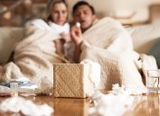 couple suffering with flu while sitting wrapped in blankets on the sofa at home, throwing tissues on coffee table