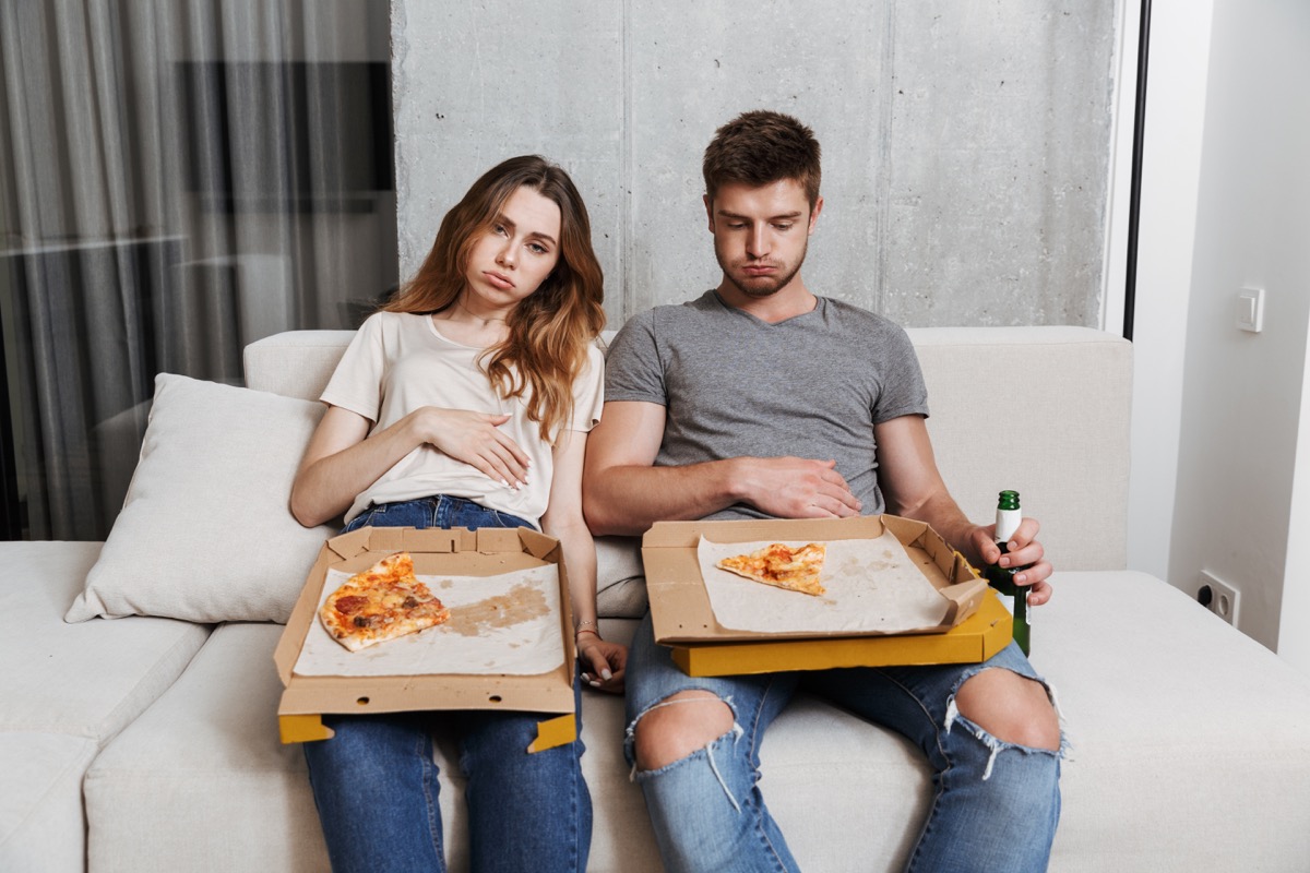 Young couple uncomfortably full from overeating pizza on couch