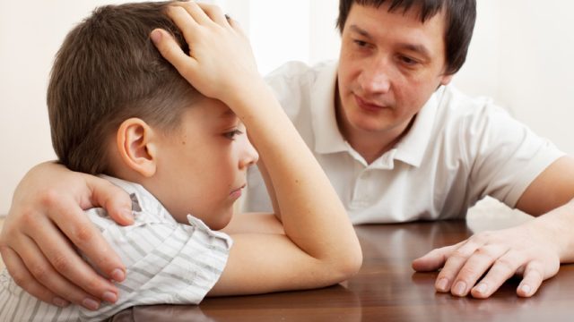 asian dad comforts young son who looks upset at table, has head in hands