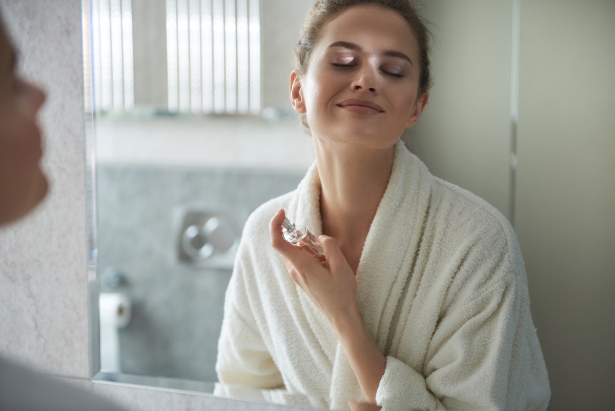 Self care concept. Waist up reflection of young smiling woman in white bathrobe enjoying her morning while applying favorite perfume before bathroom mirror (Self care concept. Waist up reflection of young smiling woman in white bathrobe enjoying her m