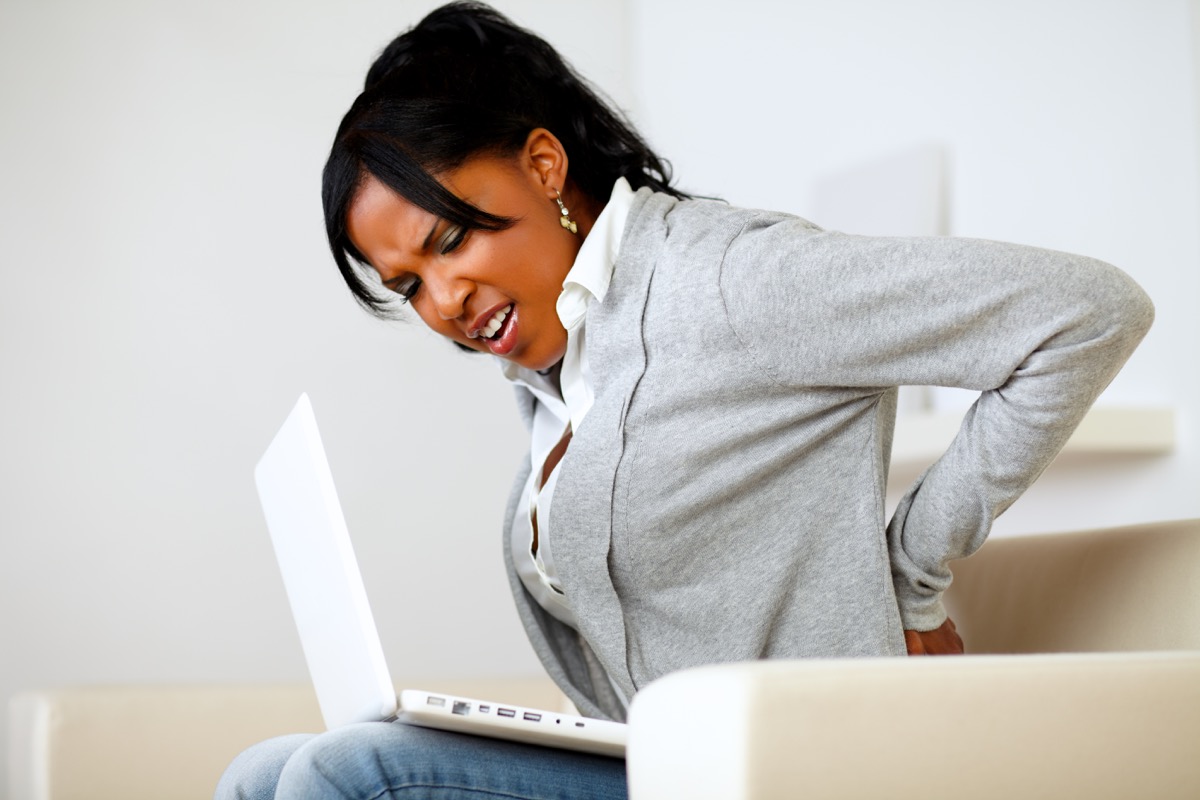 Woman experiencing back pain while sitting on a couch on her laptop
