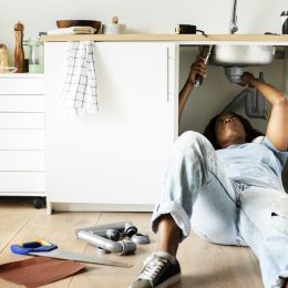 black woman laying down on floor fixing pipes under kitchen sink