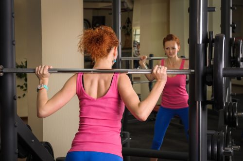 young red haired girl doing squats with barbell on smith machine in front of mirror