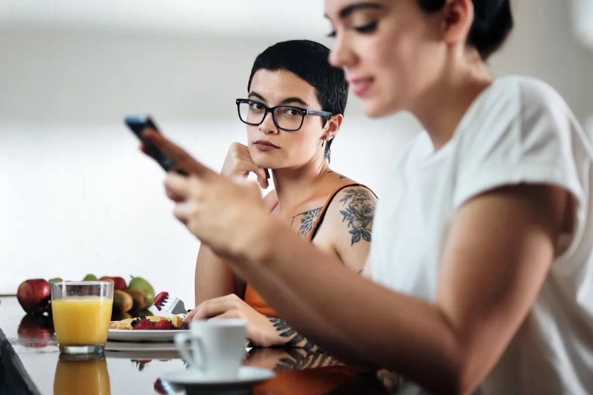 two lesbian women at home eating breakfast, partner chatting on mobile telephone. Young woman being ignored by her girlfriend and feeling jealous