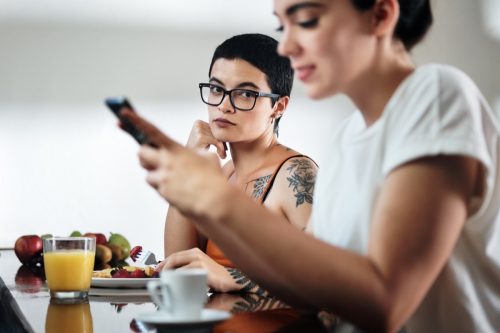 two women at home eating breakfast, partner chatting on mobile telephone. Young woman being ignored by her girlfriend and feeling jealous