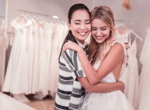 two women hugging in wedding gown and casual clothing at bridal shop