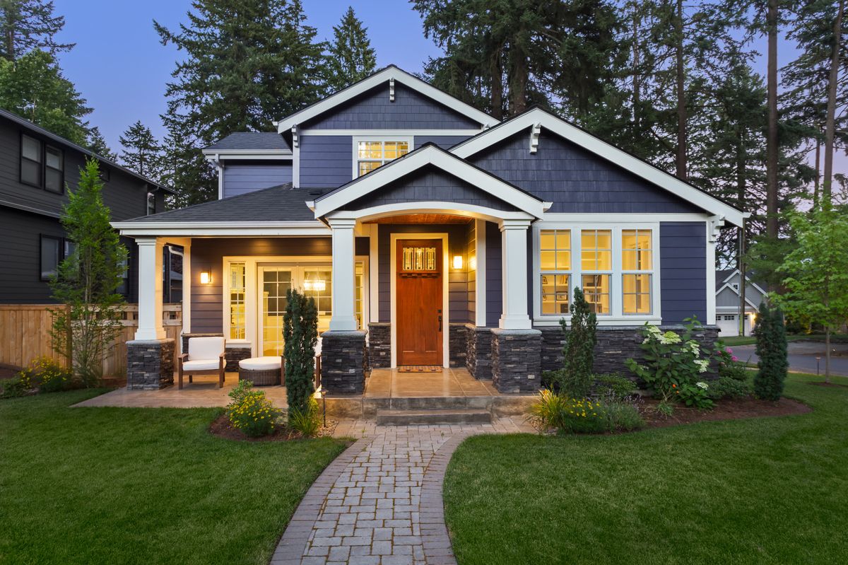 facade of dark blue home with manicured lawn, landscaping, and backdrop of trees