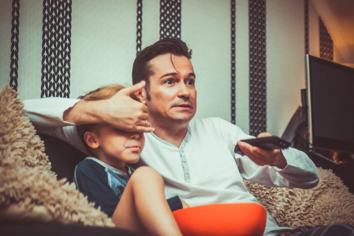 Shocked father covering son's eyes and changing channel while watching inappropriate television content at home.