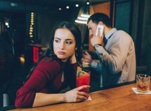 sad girl looking off while her date is on the phone at the bar
