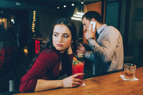 sad girl looking off while her date is on the phone at the bar