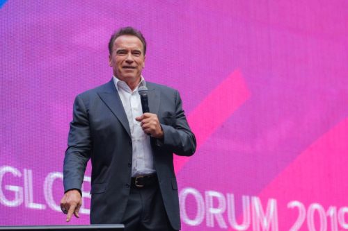 arnold schwarnegger stands on stage with pink and purple background in russia