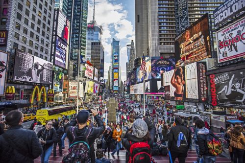 View of crowded Times Square in New York City. People are at major commercial intersection and neighborhood in Midtown Manhattan. Commercial signs are on buildings in city. Travel Locations.