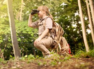 young girl scout kneeling in woods looking through binoculars