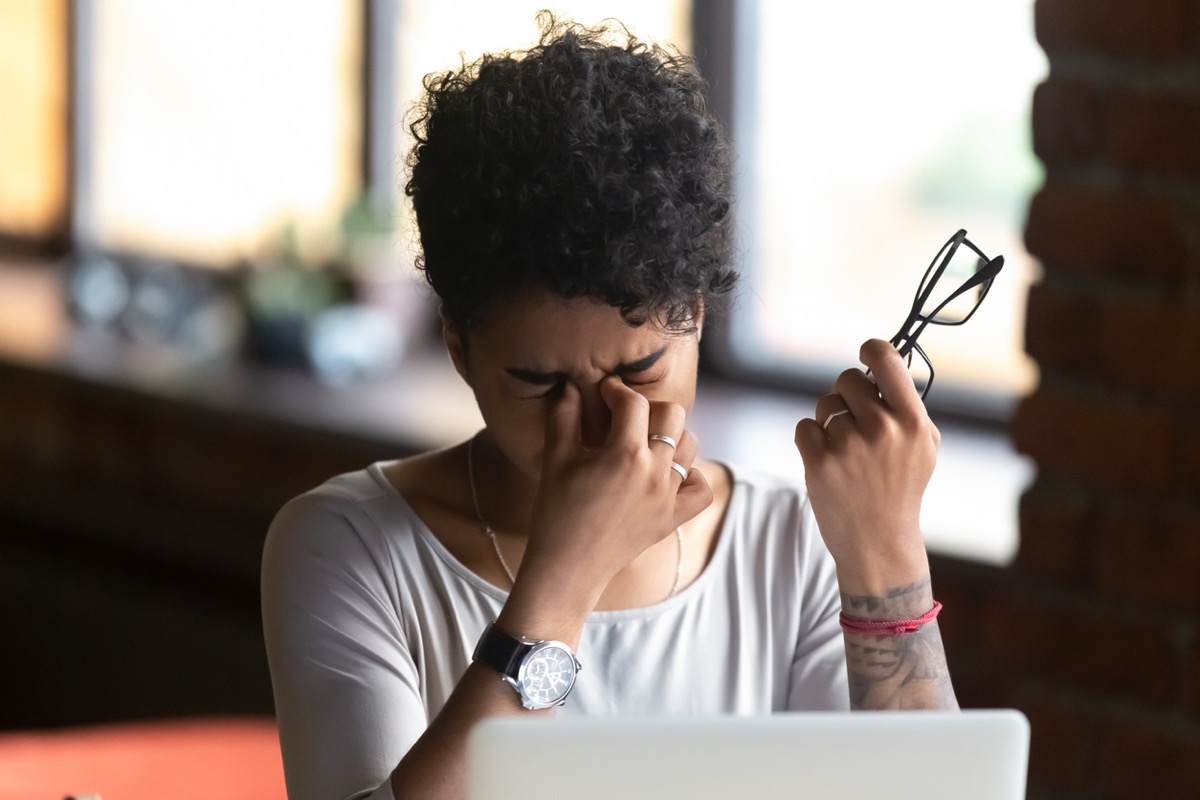 black woman rubbing her eyes and holding her glasses in front of a computer