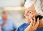 Serious senior woman answering smart phone with man in background at home