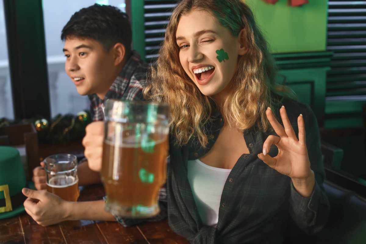 woman with green shamrock sticker on her face and beer in hand celebrating st patrick's day in a bar