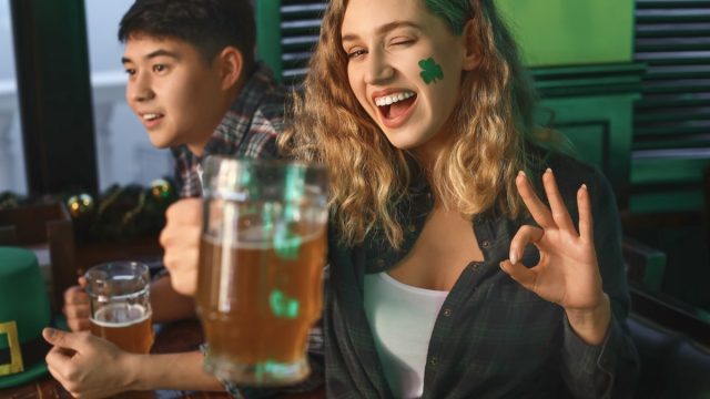 woman with green shamrock sticker on her face and beer in hand celebrating st patrick's day in a bar